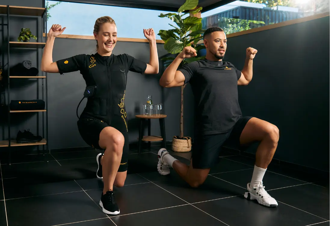 Ultymatywny personalny trening EMS – Trenuj indywidualnie z SYMBIONT EMS Kobieta w stroju EMS wykonuje ćwiczenia pod okiem profesjonalnego trenera, aby zapewnić skuteczny i indywidualny trening. Trenuje w minimalistycznie urządzonym, jasnym pomieszczeniu.