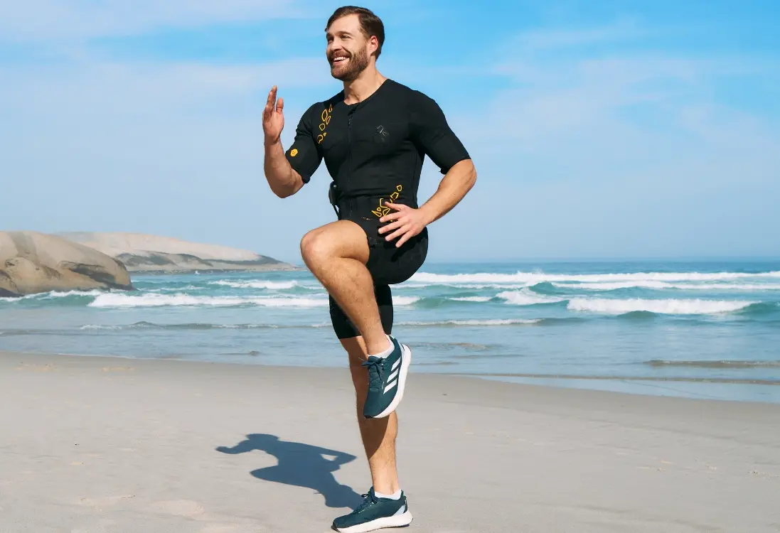 Mężczyzna w stroju EMS trenuje na plaży, wykonując skuteczny i intensywny trening, aby zredukować tkankę tłuszczową i wspierać odchudzanie za pomocą EMS.